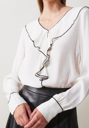 Woman wearing a white long-sleeve blouse with ruffled black-edged collar and cuffs, paired with a high-waisted black leather skirt.