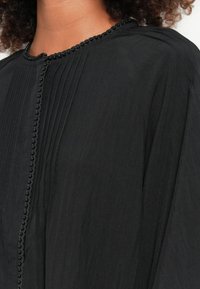 Blusa preta com pregas, decote arredondado e borda decorativa com contas na parte da frente. Feita de um tecido leve e texturizado.