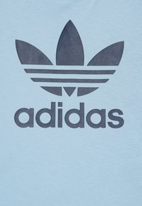 Svetlo modré tričko s veľkým tmavomodrým logom Adidas, ktoré obsahuje tri listovité tvary a tri horizontálne pruhy pod textom.