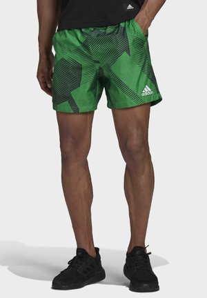 Mann trägt grüne Adidas-Shorts mit schwarzen geometrischen Mustern und schwarze Sneaker, steht mit einer Hand in der Tasche.
