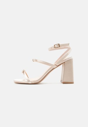 Beige Sandalette mit hohem Blockabsatz, dünnem Zehenriemen mit einer kleinen Schleife und einem Knöchelriemen mit goldener Schnalle.