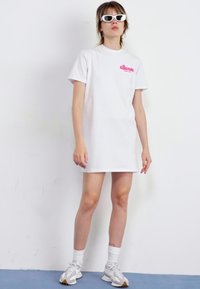 Rochie albă supradimensionată cu mâneci scurte, având un logo roz. Asortată cu ochelari de soare albi și teniși gri. Material neted, stil lejer.