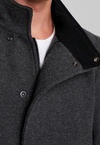 Cappotto grigio in misto lana con colletto alto, caratterizzato da dettagli in maglia nera, chiusure a bottoni a pressione e una cerniera visibile. Dettaglio del tessuto testurizzato.