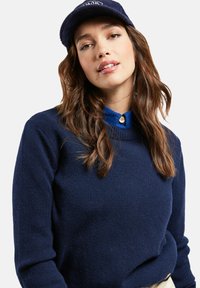 Pull en laine bleu marine avec un col rond, présentant une finition texturée et des manches longues, superposé sur une chemise bleue à col avec un bouton.