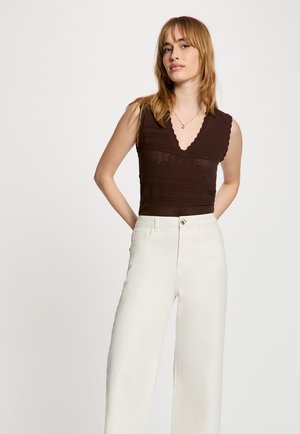 Vrouw met lichtbruin haar, die een mouwloos donkerbruin V-hals topje en witte, hooggesneden brede broek draagt, staand met de handen achter de rug.