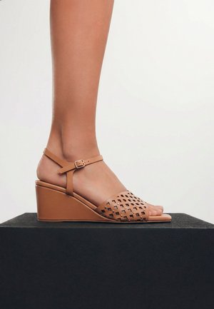 Pie con sandalia de cuña de cuero color beige con tira perforada en el dedo y hebilla en el tobillo, de pie sobre una plataforma negra sobre fondo blanco.