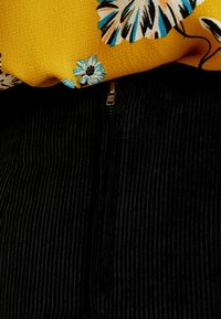Top amarillo con estampado floral, superpuesto sobre material de pana negra con acanaladuras verticales; presenta una cremallera dorada visible en el centro.