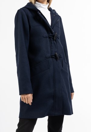 Marineblå duffelcoat med hætte og træknapper, med påsyede lommer, båret over en hvid rullekrave og sorte bukser på en stående person.