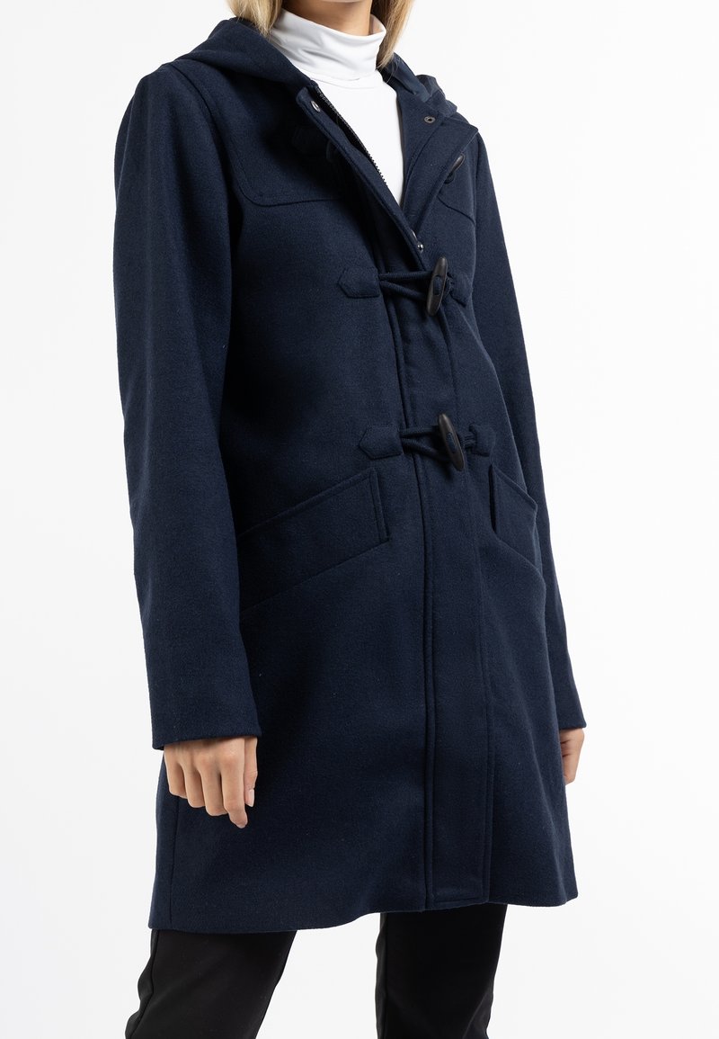 Manteau duffle bleu marine à capuche avec fermetures à boutons à bascule, poches plaquées, porté sur un col roulé blanc et un pantalon noir sur une personne debout.