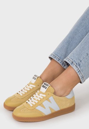 Zapatillas amarillas y azul claro con cordones blancos, usadas con vaqueros azul claro remangados sobre un fondo blanco.