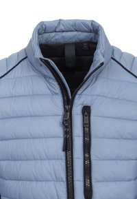 Világoskék párnázott mellény álló gallérral, elülső cipzárral és fekete díszítéssel. Jellemzője a vízszintes varrás és a cipzáros zseb.