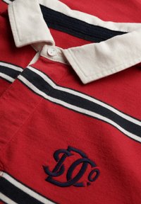 Rød polo-shirt med hvide og marineblå striber, en cremefarvet krave og et broderet logo i marineblå på brystet. I bomuldsstof med struktureret overflade.