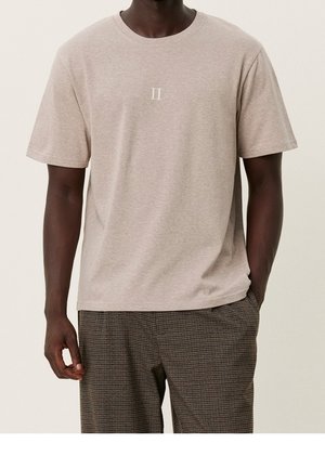 Homme portant un t-shirt beige à manches courtes avec le logo "II" sur la poitrine et un pantalon à carreaux marron, debout devant un fond uni.