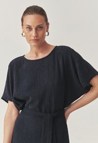 Una donna con i capelli tirati indietro indossa orecchini a cerchio d'oro e un vestito nero lavorato con una cintura in vita, mentre posa su uno sfondo chiaro e semplice.