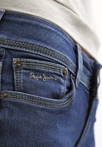 Blå denimjeans har en bakficka med ett sydd logotyp, bronsfärgad hårdvara och kontrasterande gul sömnad längs sömmarna.