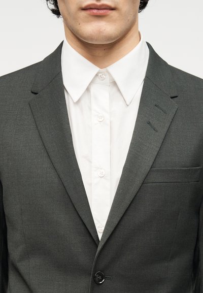 Σκούρο γκρι σακάκι με ματ φινίρισμα, κούμπωμα με ένα κουμπί και πέτο με χαραγή, συνδυασμένο με λευκό πουκάμισο με κουμπιά.