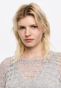 Top de punto en gris claro, con un patrón de tejido abierto, escote redondo y ribetes acanalados. La modelo lleva pendientes de oro; su cabello está despeinado.
