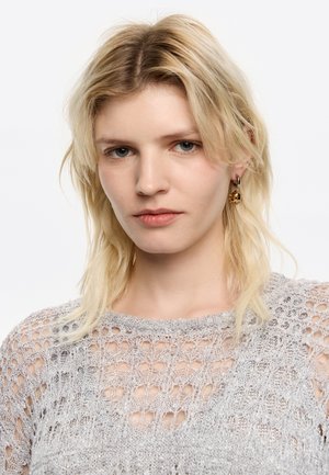 Top grigio chiaro a maglia con un motivo a rete aperta, scollatura rotonda e orli a coste. Il modello indossa orecchini dorati; i capelli sono disordinati.