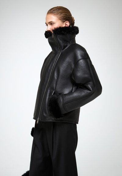 Giacca di pelle nera con colletto alto, dotata di dettagli in pelliccia sintetica sul colletto e sui polsini; texture liscia, design corto e chiusura con zip.
