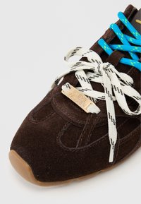 Sapatilha de camurça castanha com atacadores brancos e azuis em padrão, apresentando uma etiqueta de metal dourada com a inscrição "STEVE MADDEN" e uma sola de borracha.