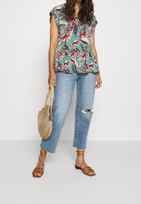 Grøn top med blomsterprint og korte ærmer, parret med lyseblå ripped jeans og brune sandaler med stropper, mens jeg holder en rund stråtaske.