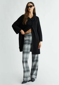 Černý chunky pletený cardigan s límcem, doplněný šedými kostkovanými kalhotami s rozšířenými nohavicemi. Má texturovanou látku a volný střih.