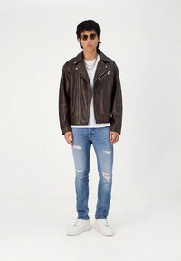 Brun skinn motorsykkeljakke med sølvfargede detaljer, brukt over en hvit t-skjorte. Kombinert med slitte blå jeans og hvite joggesko.