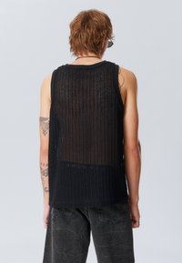 Top noir sans manches avec un design texturé et semi-transparent à rayures verticales. Tatouages notables visibles sur le bras.