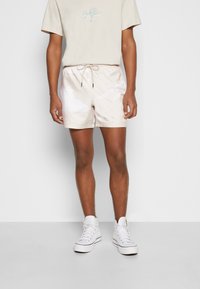 Vaaleanbeige shortsit, joissa on sileä pinta, joustava vyötärö, nyöriyksityiskohta ja hillityt valkoiset kuviot. Yhdistetty valkoisiin tennareihin.