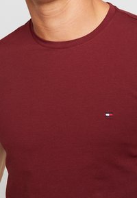 Camiseta de algodón color burdeos con cuello redondo. Presenta un pequeño logo bordado con un diseño en rojo, blanco y azul en el pecho.