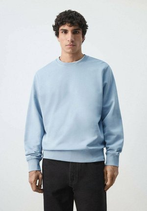 Joven con cabello rizado que lleva una sudadera azul claro de cuello redondo y pantalones negros, de pie frente a un fondo claro y liso.