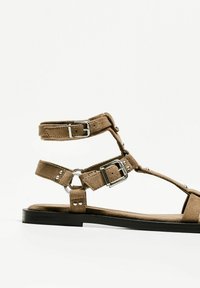 Bruna mockaskinnsandaler med flera remmar, silverfärgade detaljer, en platt svart sula och cirkulär spänne.