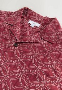 Lyhythihainen viininpunainen paita, joka on valmistettu pehmeästä kankaasta ja jossa on valkoinen geometrinen kuvio, napitus ja levitetty kaulus. Brändin etiketti näkyvissä.