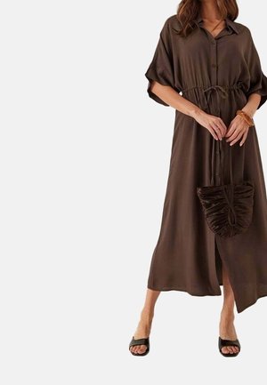 Femme portant une robe midi marron à boutons avec des manches courtes, tenant un sac à main froncé assorti et portant des talons ouverts noirs.