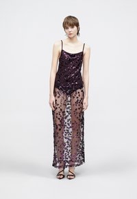 Haut orné de sequins violets avec fines bretelles, associé à une jupe longue transparente ornée de motifs floraux violets et noirs. Sandales noires à lanières.