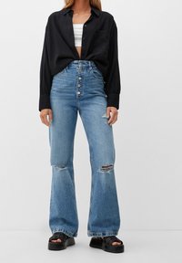 Jeans strappati in denim chiaro a vita alta, con un taglio wide-leg, abbinati a una camicia nera con bottoni sopra un top corto bianco e sandali neri con plateau.