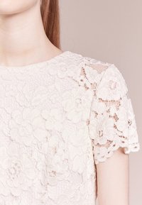 Kvinna som bär en vit spetsblus med kort ärm och blommönster, stående mot en enfärgad ljusbeige bakgrund.