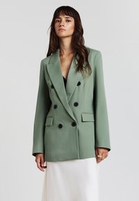 Blazer vert à double boutonnage en tissu léger, arborant des boutons noirs, des revers à cran et des poches avant. Texture lisse et coupe décontractée.