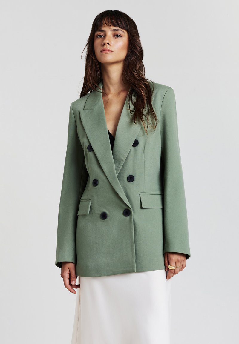 Blazer vert à double boutonnage en tissu léger, arborant des boutons noirs, des revers à cran et des poches avant. Texture lisse et coupe décontractée.