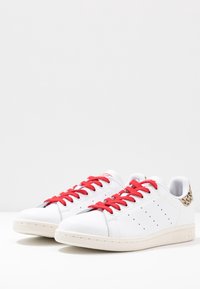 Weiße Sneakers mit roten Schnürsenkeln, die ein Obermaterial aus glattem Leder und Belüftungsöffnungen aufweisen. Auffälliger Leopardenmusterakzent an der Ferse und Gummisohle.