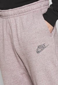 Persoon die lichtpaarse trainingsbroek draagt met glinsterend Nike-logo, elastische tailleband en hand in de rechterzak, gecombineerd met een zwart topje aan de mouw.