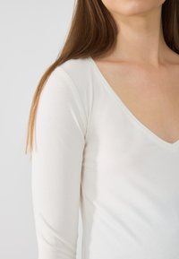 Weißes Langarmhemd mit tiefem V-Ausschnitt und weicher Textur, das durch seinen figurbetonten Schnitt eine elegante Silhouette betont.