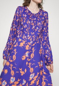 Vestido roxo com um padrão floral em toda a sua extensão, em laranja e azul, com mangas bufantes longas e decote franzido. Textura de tecido suave.