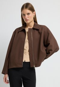 Brun zip-up jakke med en boksformet silhuet, der har en flad krave og struktureret stof. Sammen med en beige bluse og sorte bukser.