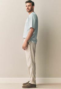 Lyseblå kortærmet t-shirt med frontlomme, parret med beige linnedbukser og brune ruskinds sko. Stående profilvisning.