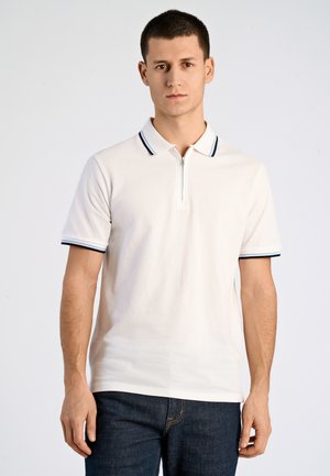 Jonge man staand, gekleed in een wit poloshirt met korte mouwen en rits, en donkere jeans, tegen een effen lichte achtergrond.