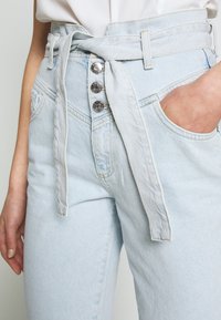 Ljusblå högmidjade jeans i denim, med ett knutet skärp, tre metallknappar och framfickor. Mjuk textur.