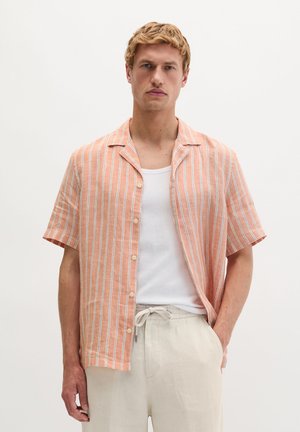 Blonde man die een perzikkleurig en wit verticaal gestreept shirt met korte mouwen draagt over een wit hemdje met beige broek met trekkoord.