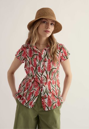 Vrouw met een bruine geweven hoed, een kort mouwens overhemd met rode en groene bladpatronen en olijfgroene broek, handen in de zakken.