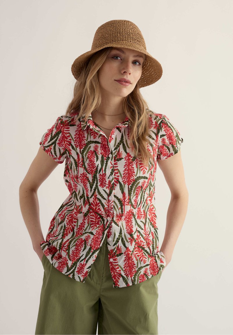 Femme portant un chapeau marron tissé, une chemise à manches courtes à boutons avec des motifs de feuilles rouges et vertes, et un pantalon vert olive, mains dans les poches.